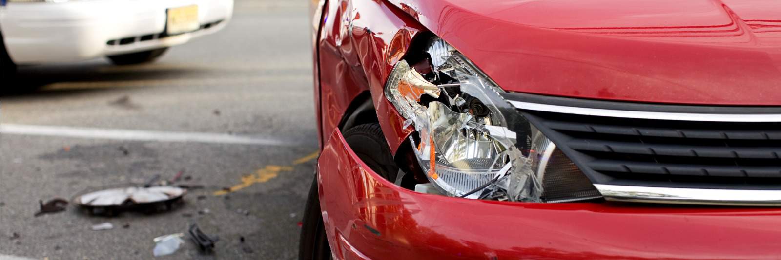 DCPD Alberta Insurance Update: A photo of a car accident on a road. Red Car with broken headlight and bent front quarter panel. Hubcap and glass is lying in the street. White car in the background.