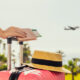 Woman with pink suitcase and passport with boarding pass standing on passengers ladder of airplane opposite sea with palm trees.