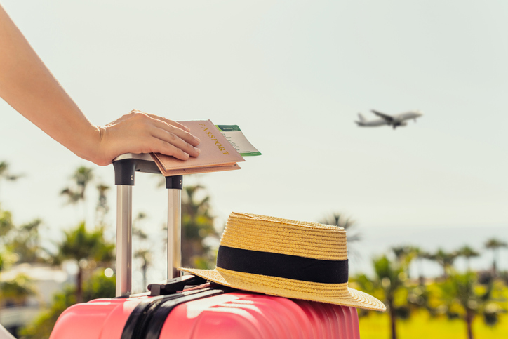 Woman with pink suitcase and passport with boarding pass standing on passengers ladder of airplane opposite sea with palm trees.